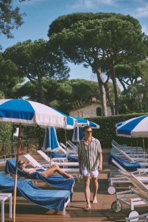Man walks on a wooden deck near lounge chairs and blue umbrellas on a sunny day, with a woman relaxing, surrounded by lush trees.