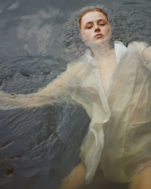 Woman in translucent shirt lying on her back in water with dark ripples and subtle waves. The portrait is serene and ethereal.
