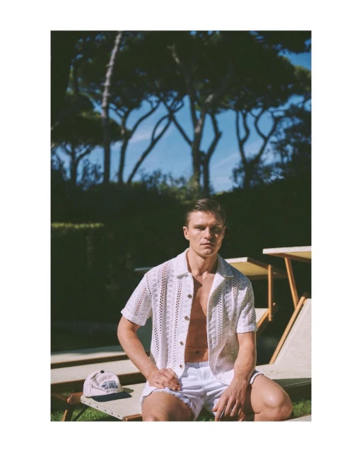 A young adult male poses outdoors wearing a knit shirt and white shorts, with a baseball cap and lawn chairs nearby under trees.
