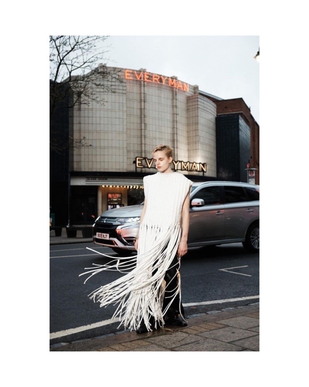 A blond model in a fringed top poses on a city sidewalk near a parked car with a theater building in the background.