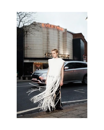 A blond model in a fringed top poses on a city sidewalk near a parked car with a theater building in the background.