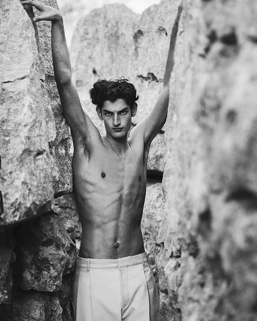 Monochrome portrait of a muscular male model posing between large rocks in natural light. The model has dark hair and light pants.