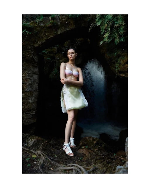 Young woman in swimwear and skirt poses in a grotto with a waterfall, creating a fashion-editorial portrait in a natural, outdoor setting.