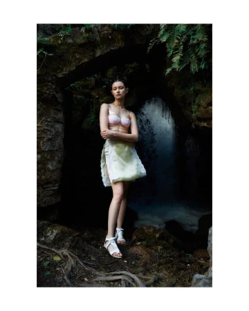 Young woman in swimwear and skirt poses in a grotto with a waterfall, creating a fashion-editorial portrait in a natural, outdoor setting.