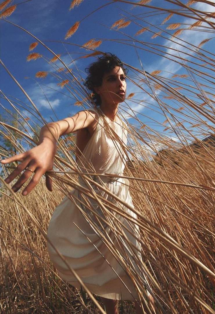 Woman in white dress stands in a field of tall, dry grass under a blue sky with light clouds, illuminated by sunlight.