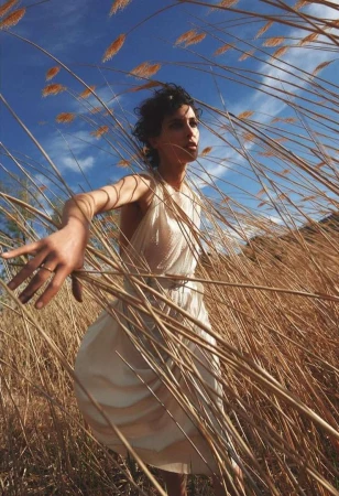 Woman in white dress stands in a field of tall, dry grass under a blue sky with light clouds, illuminated by sunlight.