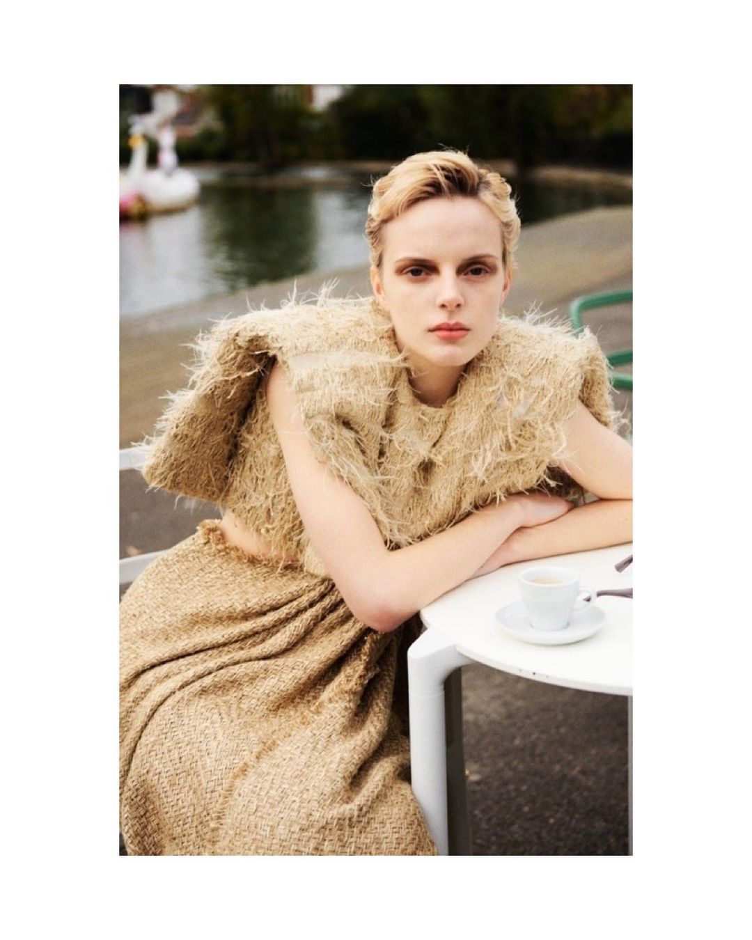 Woman with short blond hair poses in a textured tan outfit next to a round white table with a cup in an outdoor environment near water.