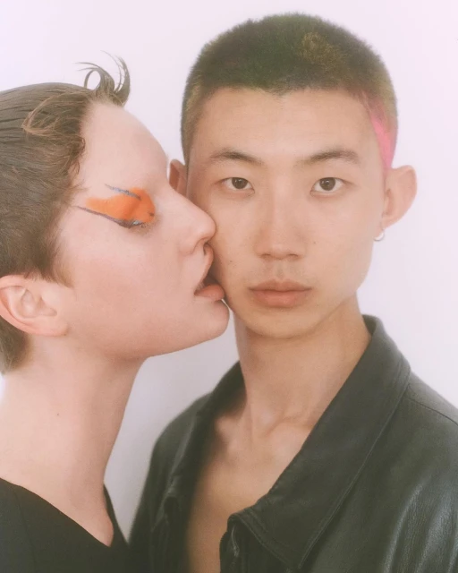 Close-up of a young adult couple. One light-skinned person with orange eye makeup leans in to another Asian person in a black jacket.