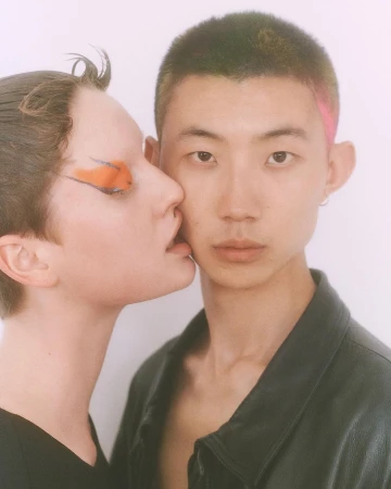 Close-up of a young adult couple. One light-skinned person with orange eye makeup leans in to another Asian person in a black jacket.