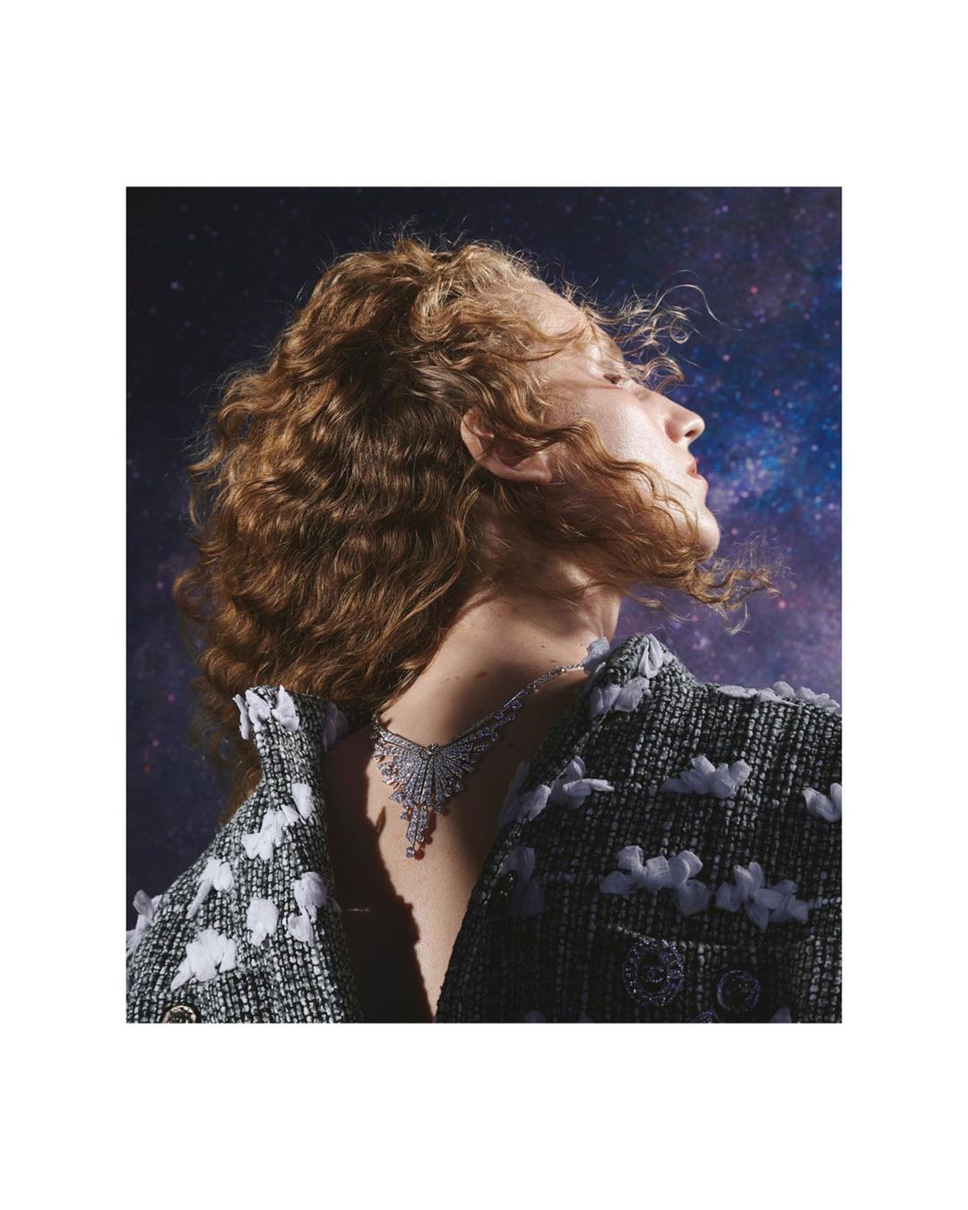 Woman in tweed jacket wearing an ornate necklace against a dark blue, starlit background in a beauty and fashion shot.