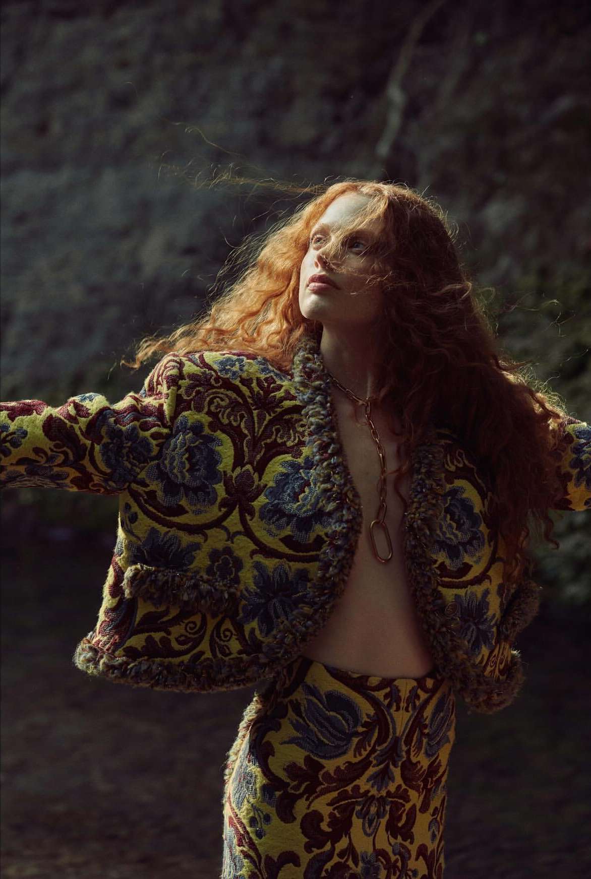 A red-haired woman in a patterned jacket and skirt poses outdoors with arms outstretched, looking upwards in a fashion editorial shot.