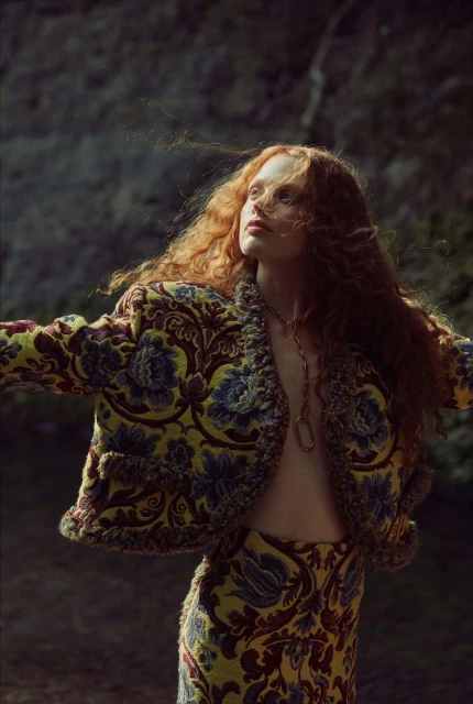 A red-haired woman in a patterned jacket and skirt poses outdoors with arms outstretched, looking upwards in a fashion editorial shot.