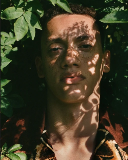 Close-up portrait of a young adult male surrounded by green foliage and dappled light, wearing a patterned shirt.
