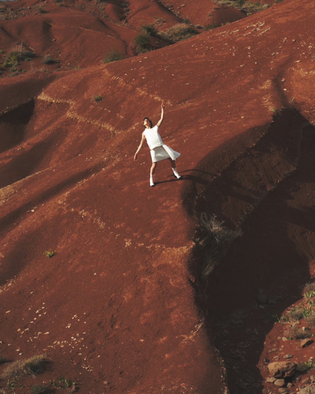 Woman in white dress poses on reddish-brown geological formations, creating contrast and striking visual composition with natural light.