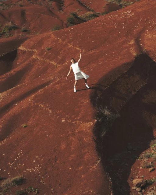 Woman in white dress poses on reddish-brown geological formations, creating contrast and striking visual composition with natural light.