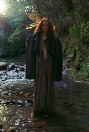 Red-haired model standing in shallow flowing river water wearing a dark structured jacket draped over shoulders and long brown dress