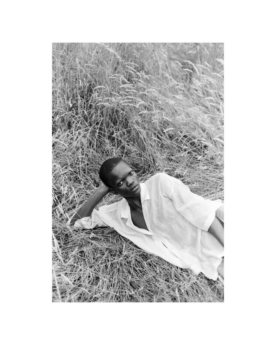 Black and white portrait of an African adult male lying in tall grass wearing a light button-down shirt, gazing directly at the camera.