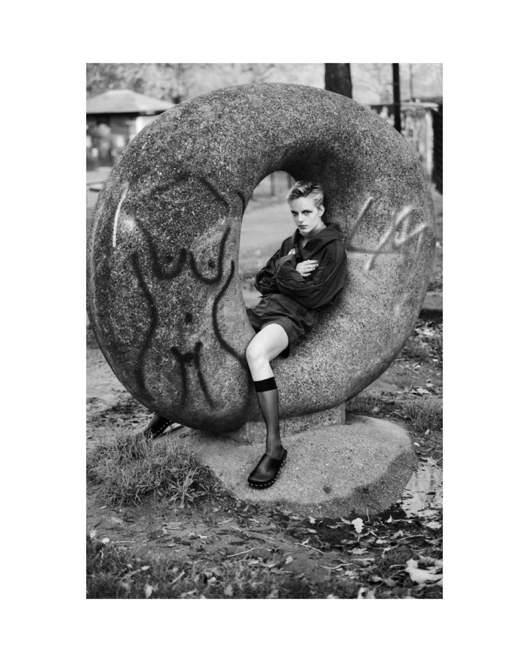 Monochrome outdoor portrait of an adult woman with short hair sitting against a large donut-shaped concrete sculpture with graffiti.