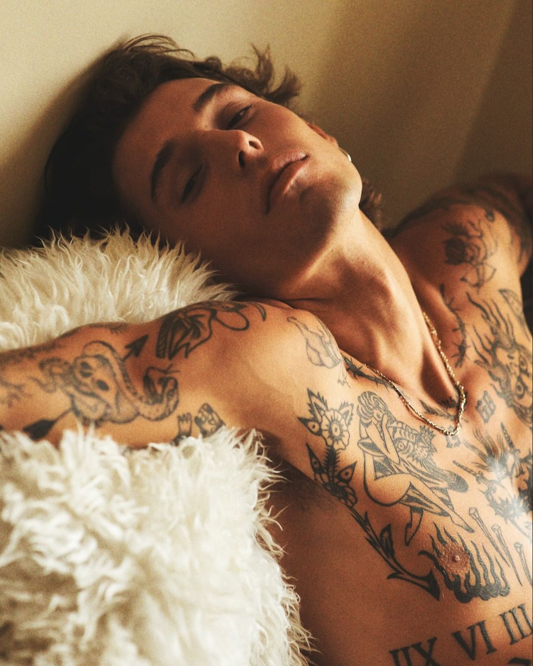 Close-up portrait of a tattooed man with dark hair resting against a fluffy white pillow in warm lighting by Olivier Yoan.