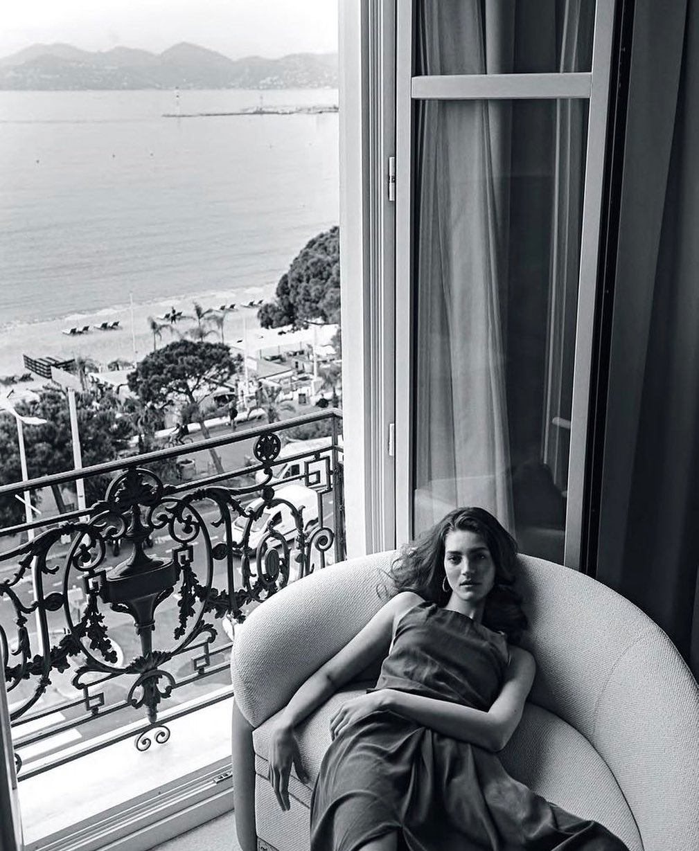 A woman lounges on a light-grey chair inside a room with an open window overlooking the ocean, beach, and city view.