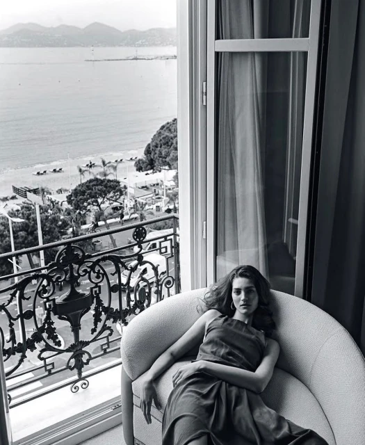A woman lounges on a light-grey chair inside a room with an open window overlooking the ocean, beach, and city view.