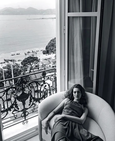 A woman lounges on a light-grey chair inside a room with an open window overlooking the ocean, beach, and city view.