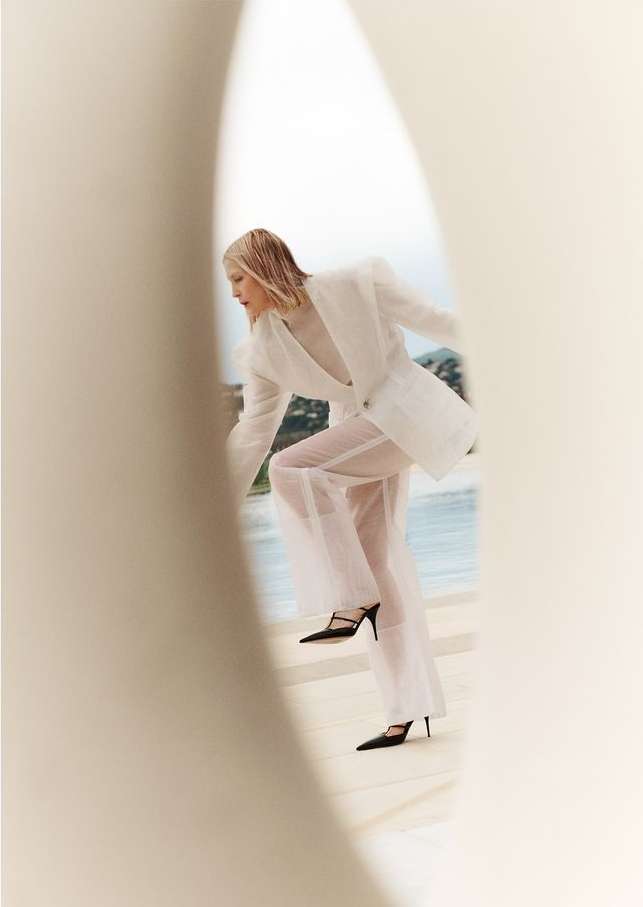 Fashion shot of a woman wearing a white blazer and sheer pants stepping onto a ledge with a sea and island view behind her.