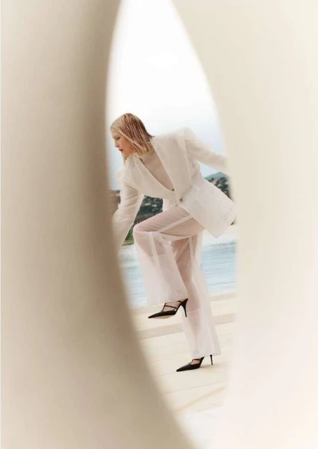 Fashion shot of a woman wearing a white blazer and sheer pants stepping onto a ledge with a sea and island view behind her.