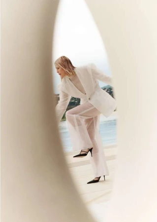 Fashion shot of a woman wearing a white blazer and sheer pants stepping onto a ledge with a sea and island view behind her.