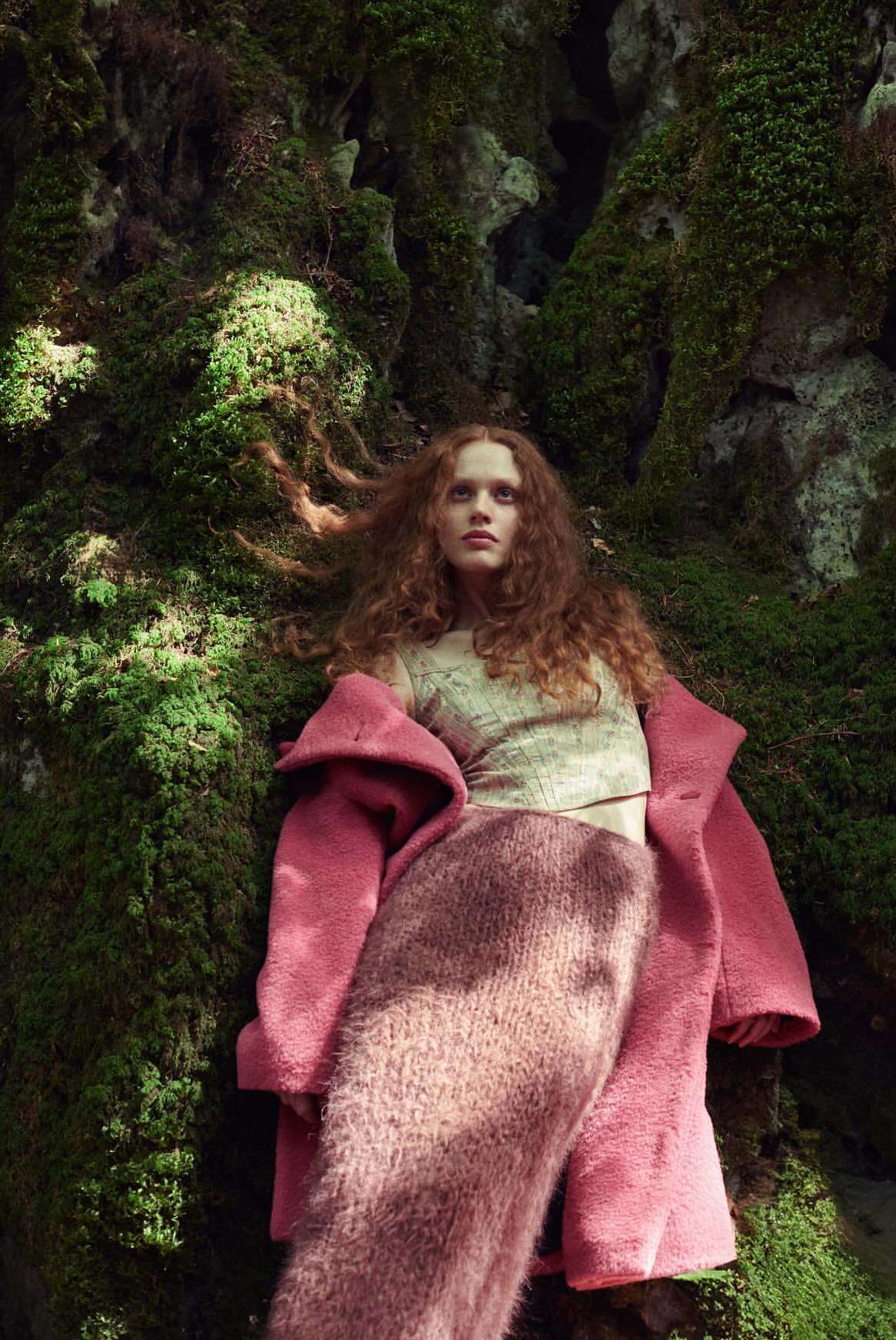 A young woman with curly red hair poses in a forest wearing a pink coat and fuzzy skirt, leaning against a mossy rock formation.