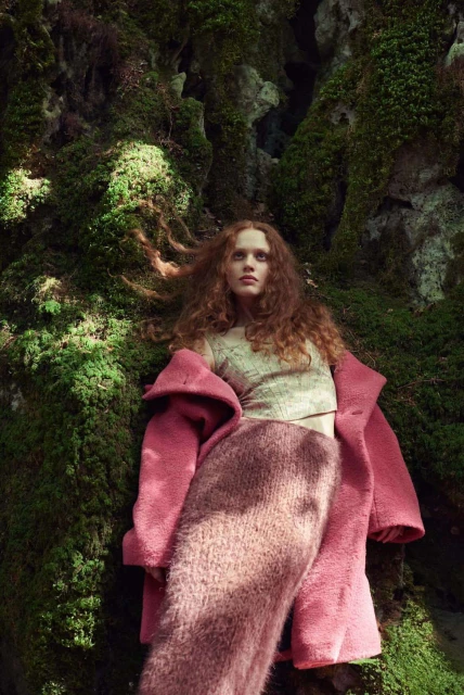 A young woman with curly red hair poses in a forest wearing a pink coat and fuzzy skirt, leaning against a mossy rock formation.