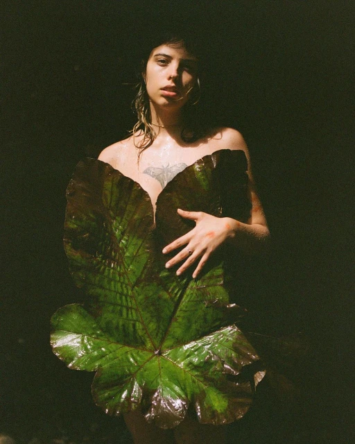 Adult woman poses with a large green leaf covering her chest in a dark environment. The image features moody lighting and natural elements.