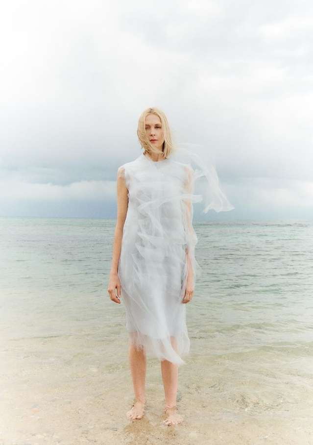Blonde woman in white dress stands in ocean surf with overcast sky. Fashion shoot at beach. Water reflects light. Wet hem and feet.