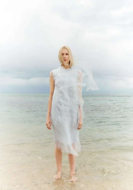 Blonde woman in white dress stands in ocean surf with overcast sky. Fashion shoot at beach. Water reflects light. Wet hem and feet.