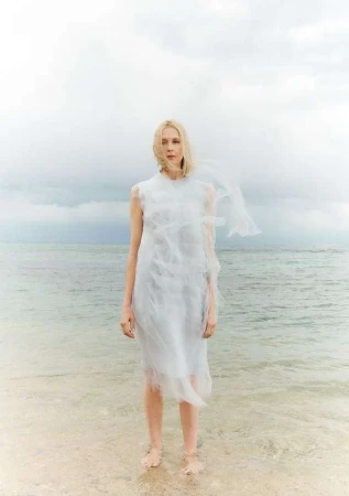Blonde woman in white dress stands in ocean surf with overcast sky. Fashion shoot at beach. Water reflects light. Wet hem and feet.