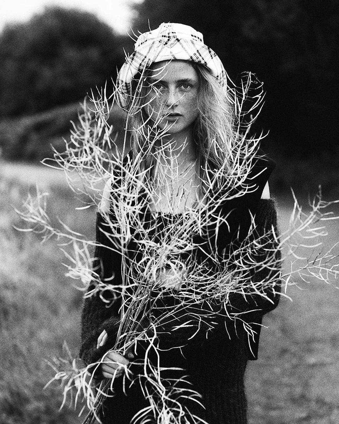 Black and white portrait of a young woman with long hair, wearing a patterned headscarf, holding a bundle of dried plants outdoors.