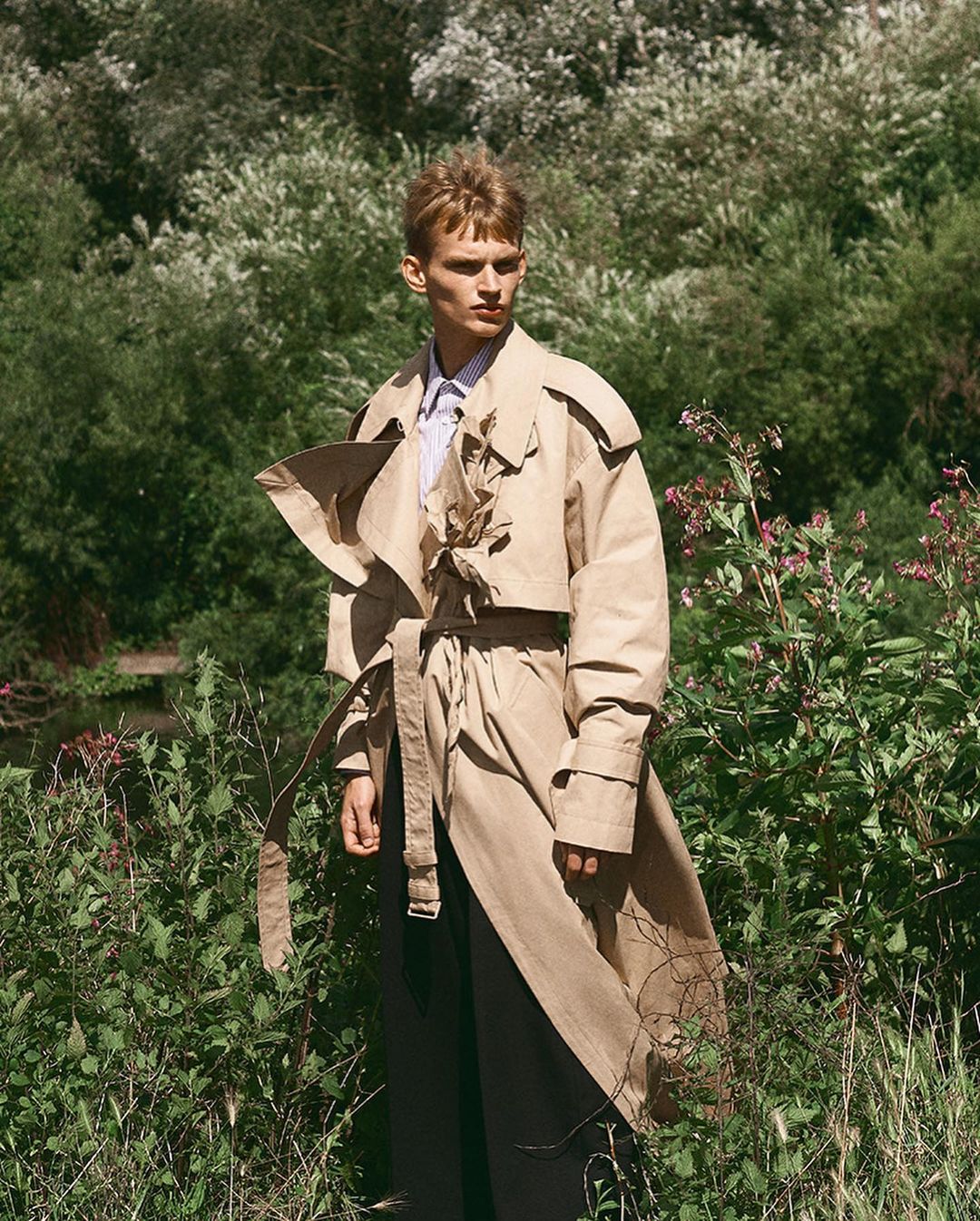 A fashionable young adult male poses outdoors in a trench coat, exuding editorial style against a lush green backdrop of plants and foliage.