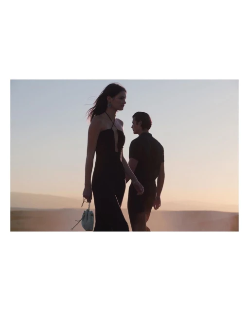 Woman in black dress with purse and man in black shirt walk outdoors at dusk against tan and light blue sky.