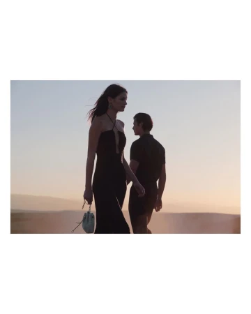 Woman in black dress with purse and man in black shirt walk outdoors at dusk against tan and light blue sky.