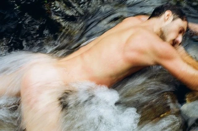 Adult male relaxing in a shallow river with a rocky riverbed and flowing water, captured with a long exposure technique.