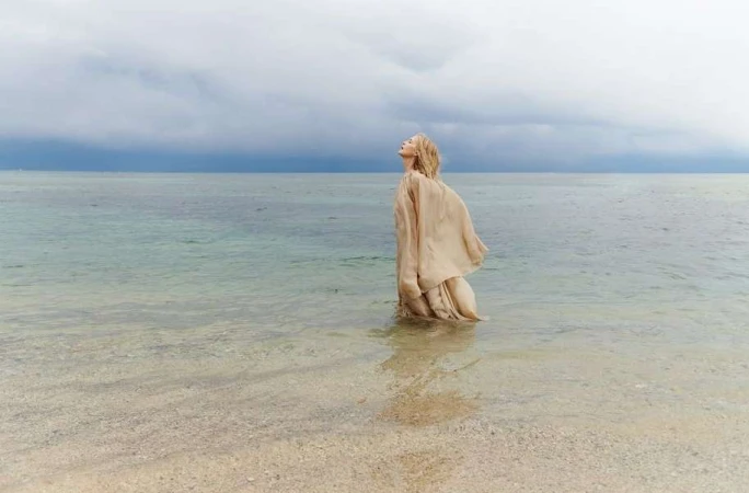 Woman in flowing beige dress standing waist-deep in ocean water under a cloudy sky