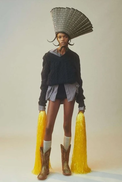 Fashion shot of a Black model in unique headwear, sweater and cowboy boots holding yellow pom-poms in a studio setting.