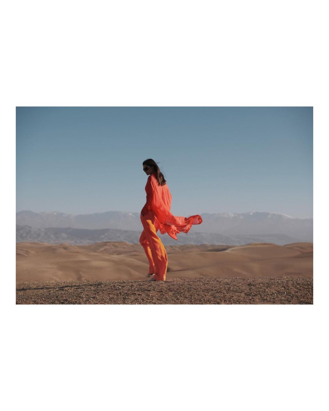 Woman in orange dress walks in desert landscape under blue sky with mountains in background, showcasing fashion in outdoor setting.