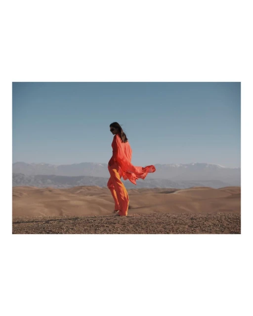 Woman in orange dress walks in desert landscape under blue sky with mountains in background, showcasing fashion in outdoor setting.