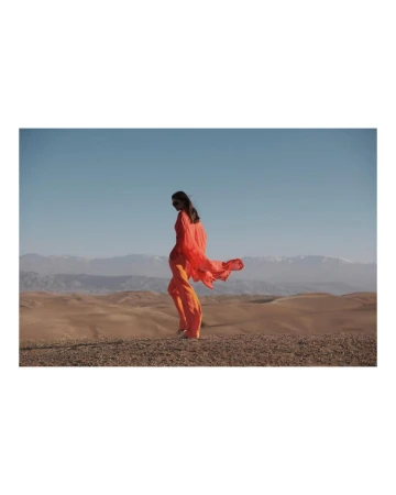Woman in orange dress walks in desert landscape under blue sky with mountains in background, showcasing fashion in outdoor setting.