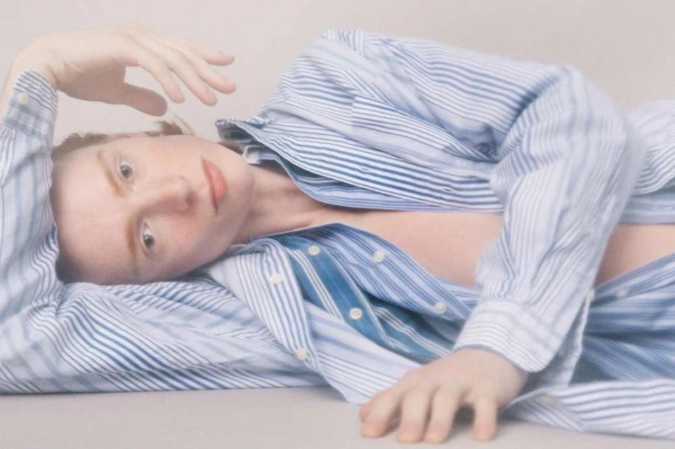 A person with freckles lies in a relaxed pose, wearing an open striped shirt against a soft, neutral-toned backdrop in a serene composition.