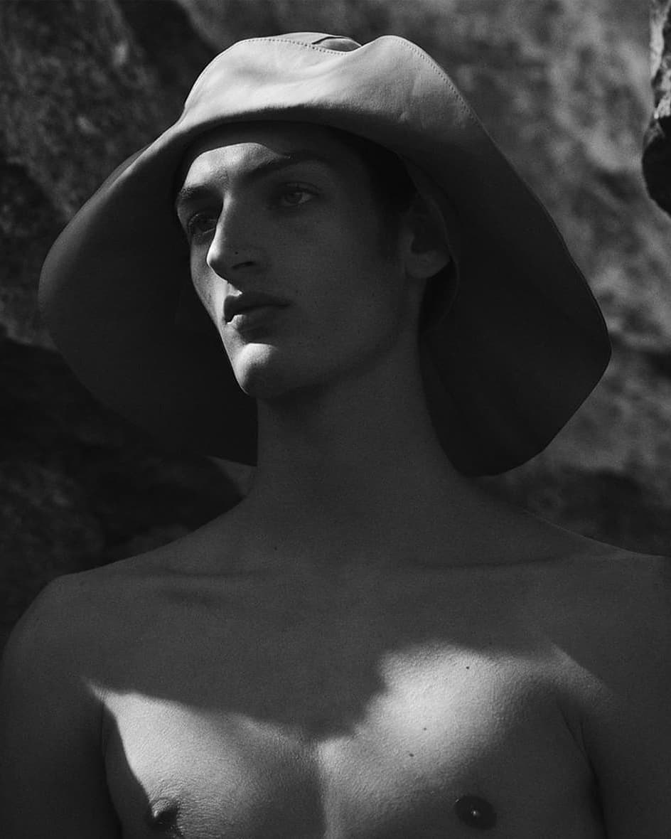 Black and white portrait of a shirtless adult male wearing a wide-brimmed hat, posing in front of a textured rock wall.