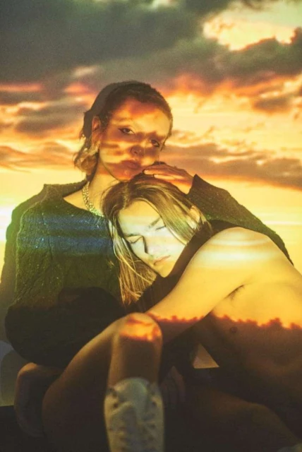 Two women embrace in a warm-toned studio portrait, with a vibrant sunset projection in the background, creating a soft, intimate atmosphere.