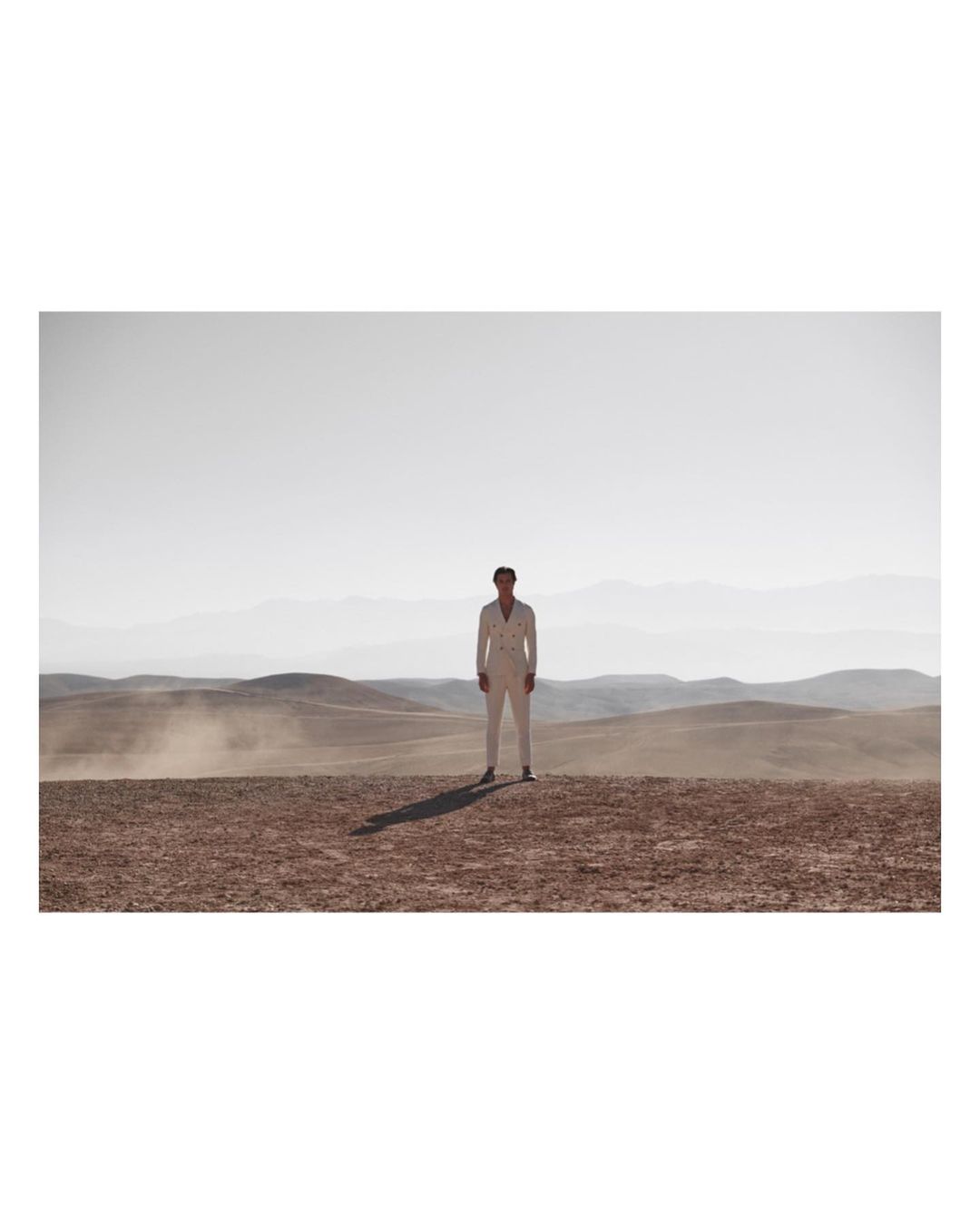 Full body shot of an adult male in white suit standing on arid landscape with distant mountains, under bright daylight.