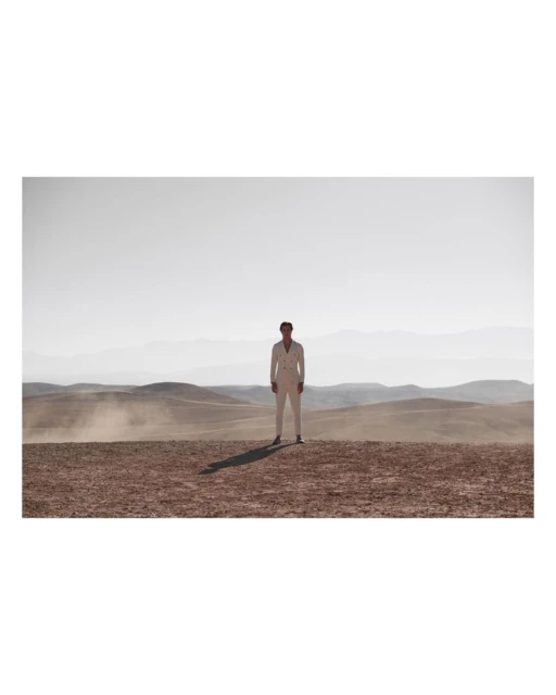 Full body shot of an adult male in white suit standing on arid landscape with distant mountains, under bright daylight.